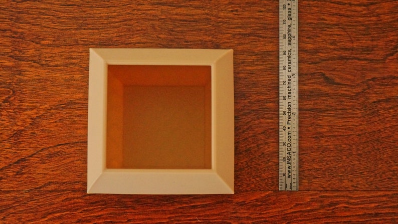 May include: A square, off-white shadow box with an orange interior sits on a wood-grain surface. A metal ruler is positioned next to the box. The shadow box is approximately 4 inches wide.