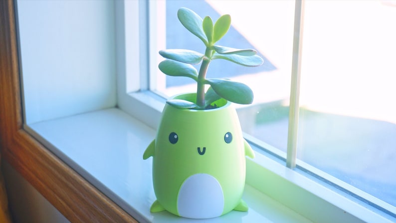 May include: A green ceramic planter shaped like a cartoon character with a smiling face. The planter has a white belly and is holding a small green succulent plant.