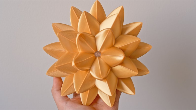May include: A gold-colored, geometric, 3D printed flower with a starburst design. The flower is made of many small, overlapping petals.
