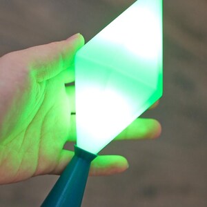 May include: A teal-colored, cone-shaped base supports a glowing, green, diamond-shaped light. The light is held in a hand against a blurred background. The light appears to be emitting a bright, even glow.