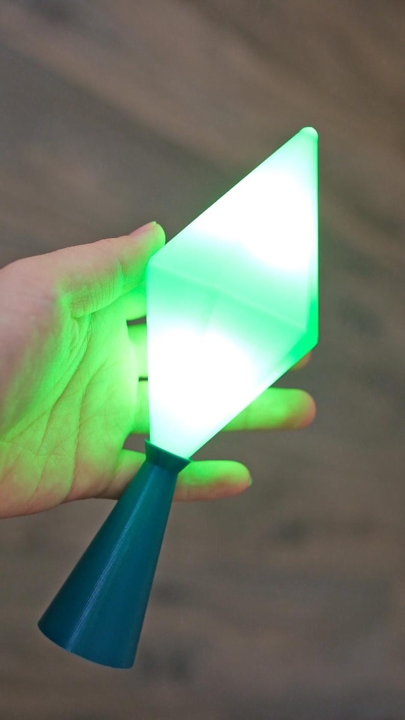 May include: A teal-colored, cone-shaped base supports a glowing, green, diamond-shaped light. The light is held in a hand against a blurred background. The light appears to be emitting a bright, even glow.