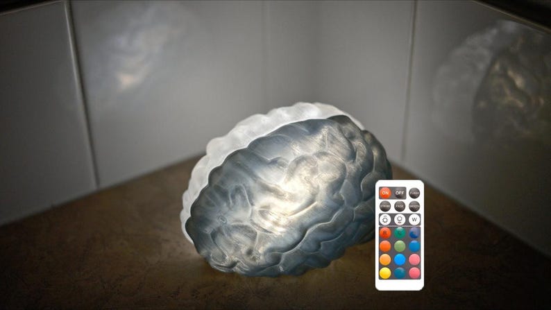 Pu&ograve; includere: Una lampada a forma di cervello con un design strutturato grigio e bianco. La lampada &egrave; illuminata e un telecomando con opzioni di colore &egrave; visibile. La lampada &egrave; su una superficie marrone, con uno sfondo bianco e grigio.