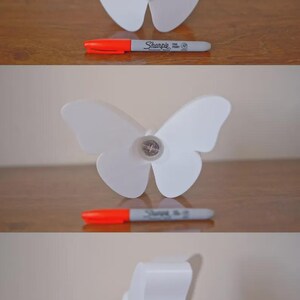 May include: A white butterfly-shaped decorative object with a Sharpie marker next to it. The marker is gray with an orange cap. The butterfly has a simple, elegant design.