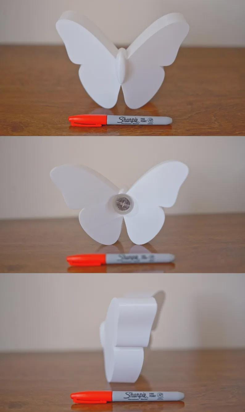 May include: A white butterfly-shaped decorative object with a Sharpie marker next to it. The marker is gray with an orange cap. The butterfly has a simple, elegant design.