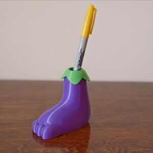 May include: Purple eggplant-shaped pen holder with a green top, holding a yellow pen.