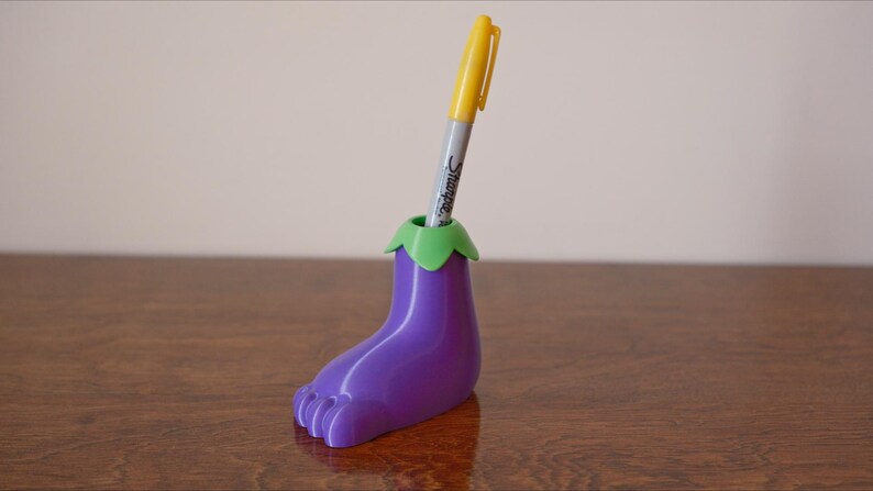 May include: Purple eggplant-shaped pen holder with a green top, holding a yellow pen.