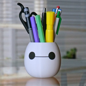 Pu&ograve; includere: Portapenne in ceramica bianca a forma di Baymax dal film Big Hero 6. Il portapenne ha una linea nera per la bocca e due cerchi neri per gli occhi. Il portapenne &egrave; pieno di varie penne e pennarelli, tra cui un pennarello viola, un pennarello verde, un evidenziatore giallo, una penna nera e una penna blu.