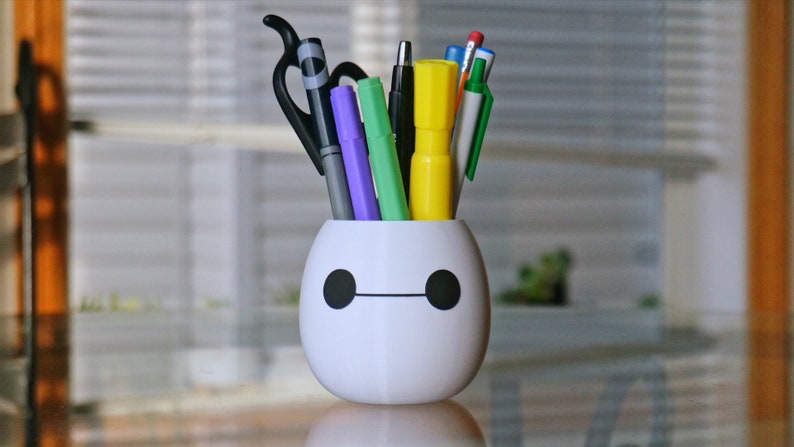 Pu&ograve; includere: Portapenne in ceramica bianca a forma di Baymax dal film Big Hero 6. Il portapenne ha una linea nera per la bocca e due cerchi neri per gli occhi. Il portapenne &egrave; pieno di varie penne e pennarelli, tra cui un pennarello viola, un pennarello verde, un evidenziatore giallo, una penna nera e una penna blu.
