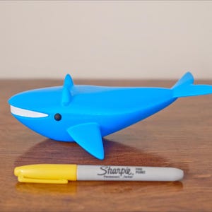 May include: A blue whale figurine with white accents and a black eye. A yellow and gray Sharpie marker is placed in front of the whale. The marker has the text "Sharpie Fine Point" printed on it.