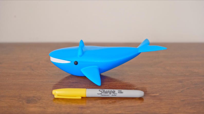 May include: A blue whale figurine with white accents and a black eye. A yellow and gray Sharpie marker is placed in front of the whale. The marker has the text "Sharpie Fine Point" printed on it.