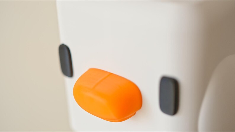 Pu&ograve; includere: Un oggetto in ceramica bianca con un design nero e arancione. L'oggetto ha due cerchi neri e un becco arancione arrotondato.