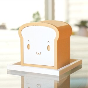 K&ouml;nnte beinhalten: Eine wei&szlig;e Brotform-Box mit einem niedlichen Comic-Gesicht. Die Box ist aus Holz und hat eine hellbraune Oberfl&auml;che. Sie steht auf einem wei&szlig;en Tablett.