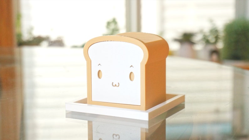 K&ouml;nnte beinhalten: Eine wei&szlig;e Brotform-Box mit einem niedlichen Comic-Gesicht. Die Box ist aus Holz und hat eine hellbraune Oberfl&auml;che. Sie steht auf einem wei&szlig;en Tablett.