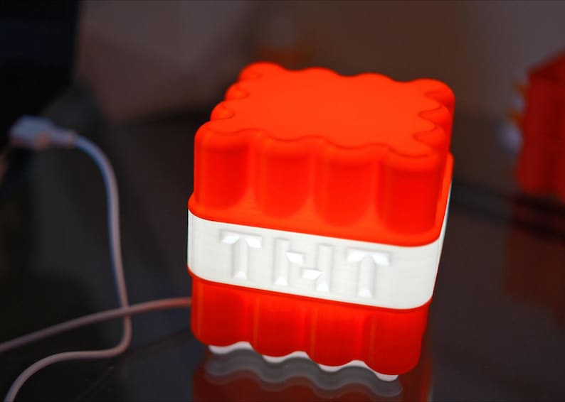 May include: A red and white Minecraft-themed night light shaped like a block of TNT. The light is plugged in and glowing.