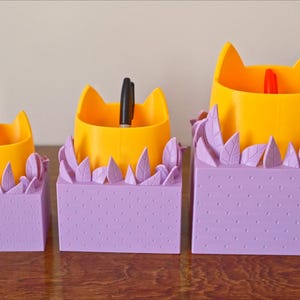 Peut inclure: Trois organiseurs de bureau en forme de chat jaune avec des bases violettes et des accents en forme de feuilles. Les organiseurs sont de tailles diff&eacute;rentes et contiennent des stylos.