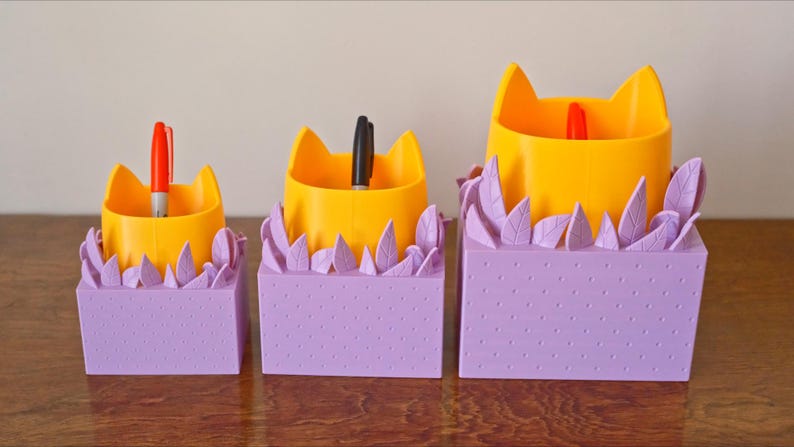 Peut inclure: Trois organiseurs de bureau en forme de chat jaune avec des bases violettes et des accents en forme de feuilles. Les organiseurs sont de tailles diff&eacute;rentes et contiennent des stylos.