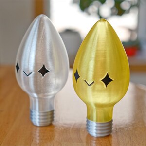 May include: Two decorative light bulb shaped objects. One is silver, the other is gold. Each has a star-shaped cutout and a small angled cutout. The base of each object is threaded, like a light bulb.