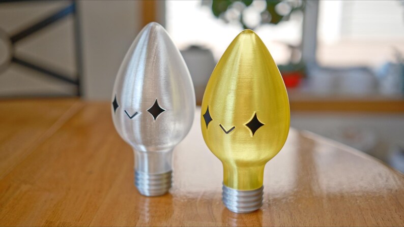 May include: Two decorative light bulb shaped objects. One is silver, the other is gold. Each has a star-shaped cutout and a small angled cutout. The base of each object is threaded, like a light bulb.