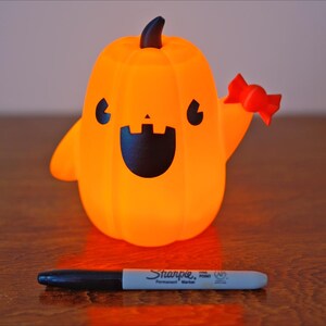 May include: A small orange pumpkin-shaped figurine with a friendly face and a red candy cane in its hand. The figurine is lit from within, giving it a warm glow.