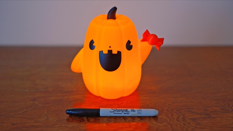 May include: A small orange pumpkin-shaped figurine with a friendly face and a red candy cane in its hand. The figurine is lit from within, giving it a warm glow.