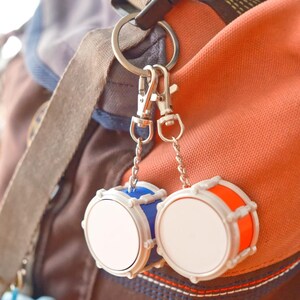 May include: A pair of miniature drum keychains, one blue and one orange, with white drumheads. The keychains are attached to a silver ring and clips, hanging against a backdrop of a backpack in shades of brown and orange.