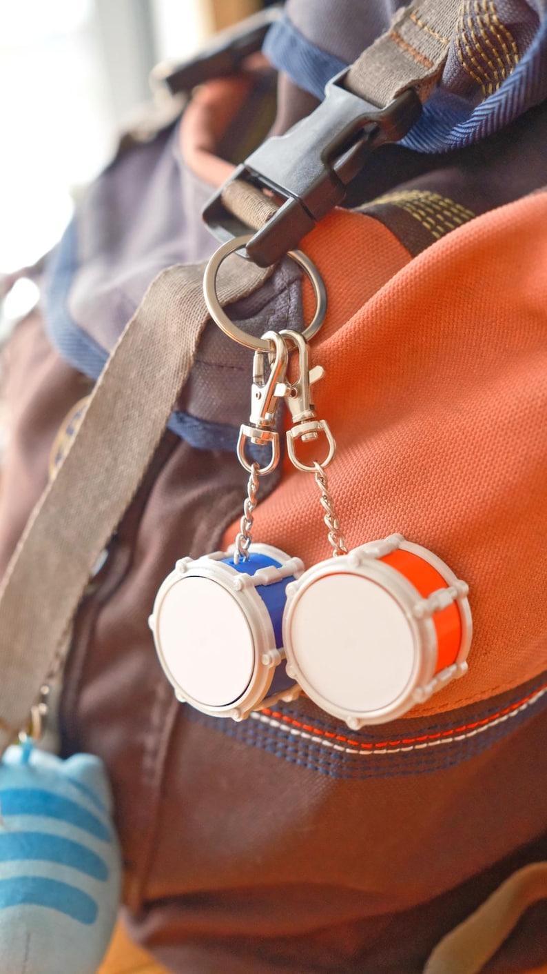 May include: A pair of miniature drum keychains, one blue and one orange, with white drumheads. The keychains are attached to a silver ring and clips, hanging against a backdrop of a backpack in shades of brown and orange.