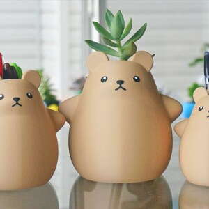 May include: Three brown bear-shaped ceramic planters, each with a different design. The first planter has a small succulent plant growing out of the top. The second planter has a bunch of colorful pens sticking out of the top. The third planter has a bunch of colorful pens and pencils sticking out of the top.