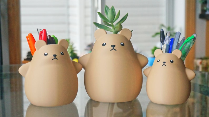 May include: Three brown bear-shaped ceramic planters, each with a different design. The first planter has a small succulent plant growing out of the top. The second planter has a bunch of colorful pens sticking out of the top. The third planter has a bunch of colorful pens and pencils sticking out of the top.