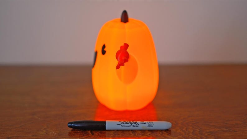 May include: Orange pumpkin-shaped Halloween decoration with a black face and red candy corn. The pumpkin is lit from within, giving it a warm glow.