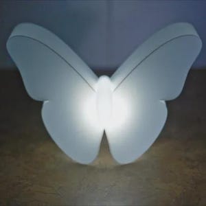 May include: A white, butterfly-shaped light fixture with a bright, glowing center. The wings are curved and symmetrical, and the fixture is set against a dark background. The light is soft and diffused.