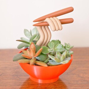 May include: A whimsical succulent arrangement in a bright orange bowl, featuring faux noodles and chopsticks. The arrangement includes various green and orange succulents, creating a playful and unique decorative piece.