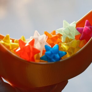 May include: A crescent-shaped orange bowl filled with colorful star-shaped objects. The stars are made of a translucent material and come in shades of red, yellow, blue, green, and white.