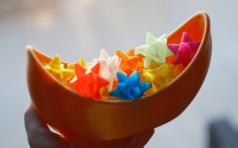 May include: A crescent-shaped orange bowl filled with colorful star-shaped objects. The stars are made of a translucent material and come in shades of red, yellow, blue, green, and white.