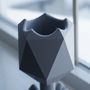 May include: A gray geometric planter with a unique, angular design. The planter is made of a smooth, matte material and has a wide opening at the top.