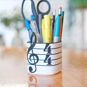 May include: A white pen holder with a musical staff design, featuring a treble clef and the numbers 4/4. The holder contains pens, pencils, scissors, and highlighters in various colors. The background is a blurred wooden table.