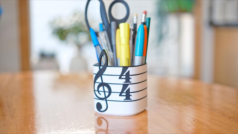 May include: A white pen holder with a musical staff design, featuring a treble clef and the numbers 4/4. The holder contains pens, pencils, scissors, and highlighters in various colors. The background is a blurred wooden table.