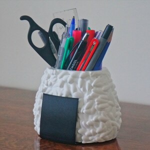 Puede incluir: Organizador de escritorio de cer&aacute;mica blanca con forma de onigiri con una pizarra negra en el lateral, lleno de bol&iacute;grafos, l&aacute;pices y un par de tijeras.