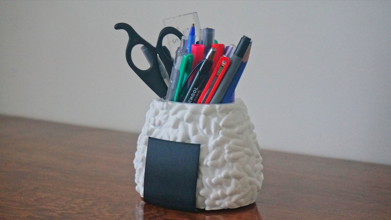 Puede incluir: Organizador de escritorio de cer&aacute;mica blanca con forma de onigiri con una pizarra negra en el lateral, lleno de bol&iacute;grafos, l&aacute;pices y un par de tijeras.