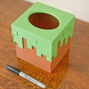A cube tissue box cover inspired by the dirt grass block from the popular video game, Minecraft, is on a wooden table with a white background and a sharpie for size. It has bright green pixelated grass at the top half and the bottom half is brown.
