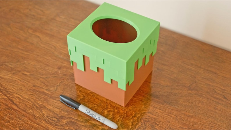 A cube tissue box cover inspired by the dirt grass block from the popular video game, Minecraft, is on a wooden table with a white background and a sharpie for size. It has bright green pixelated grass at the top half and the bottom half is brown.