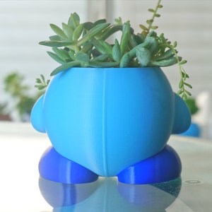 Puede incluir: Una maceta de cer&aacute;mica azul y azul claro con forma de personaje de dibujos animados con una planta suculenta en su interior.