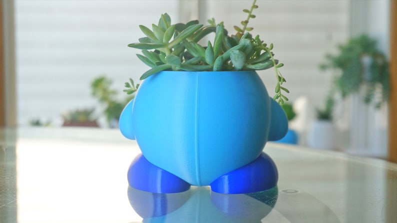 Puede incluir: Una maceta de cer&aacute;mica azul y azul claro con forma de personaje de dibujos animados con una planta suculenta en su interior.