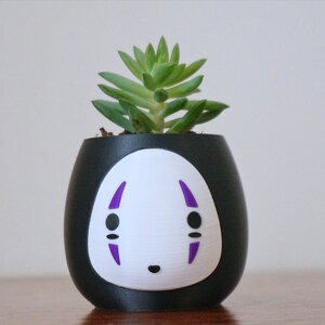 May include: A black and white ceramic planter with a No-Face design, holding a small green succulent plant. The planter has a rounded shape with a white face and purple accents. A unique decorative item.