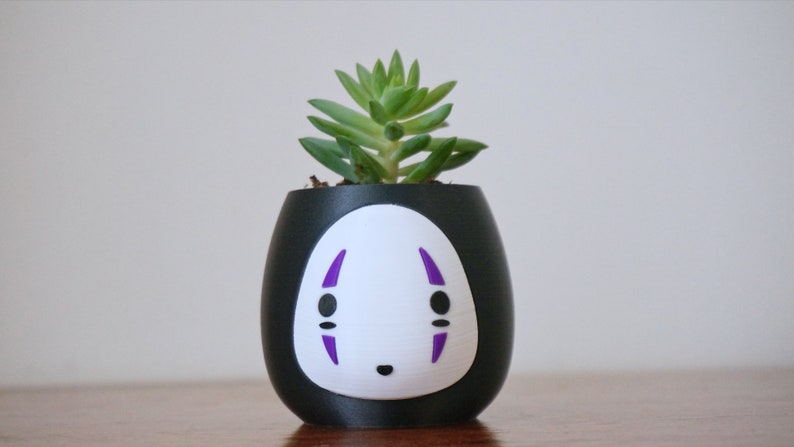 May include: A black and white ceramic planter with a No-Face design, holding a small green succulent plant. The planter has a rounded shape with a white face and purple accents. A unique decorative item.
