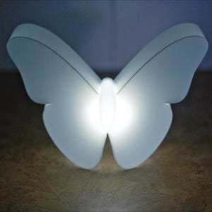 May include: A white butterfly-shaped light fixture with a bright, glowing center. The wings are softly illuminated, creating a gentle, diffused glow. The light is likely an LED or energy-efficient bulb, perfect for a bedroom or nursery.