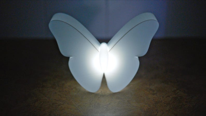 May include: A white butterfly-shaped light fixture with a bright, glowing center. The wings are softly illuminated, creating a gentle, diffused glow. The light is likely an LED or energy-efficient bulb, perfect for a bedroom or nursery.