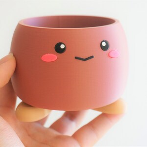 May include: A small, brown, ceramic planter with a cute cartoon face. The planter has pink cheeks, black eyes, and a small black smile. It has two small, brown feet.