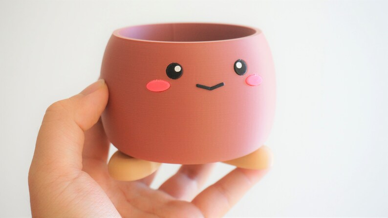 May include: A small, brown, ceramic planter with a cute cartoon face. The planter has pink cheeks, black eyes, and a small black smile. It has two small, brown feet.