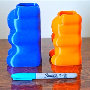 Puede incluir: Dos porta l&aacute;pices con forma de osito de goma, uno azul y otro naranja, est&aacute;n sobre una superficie de madera. Un marcador azul est&aacute; delante de ellos.