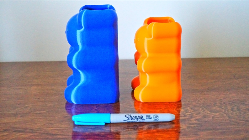 Puede incluir: Dos porta l&aacute;pices con forma de osito de goma, uno azul y otro naranja, est&aacute;n sobre una superficie de madera. Un marcador azul est&aacute; delante de ellos.